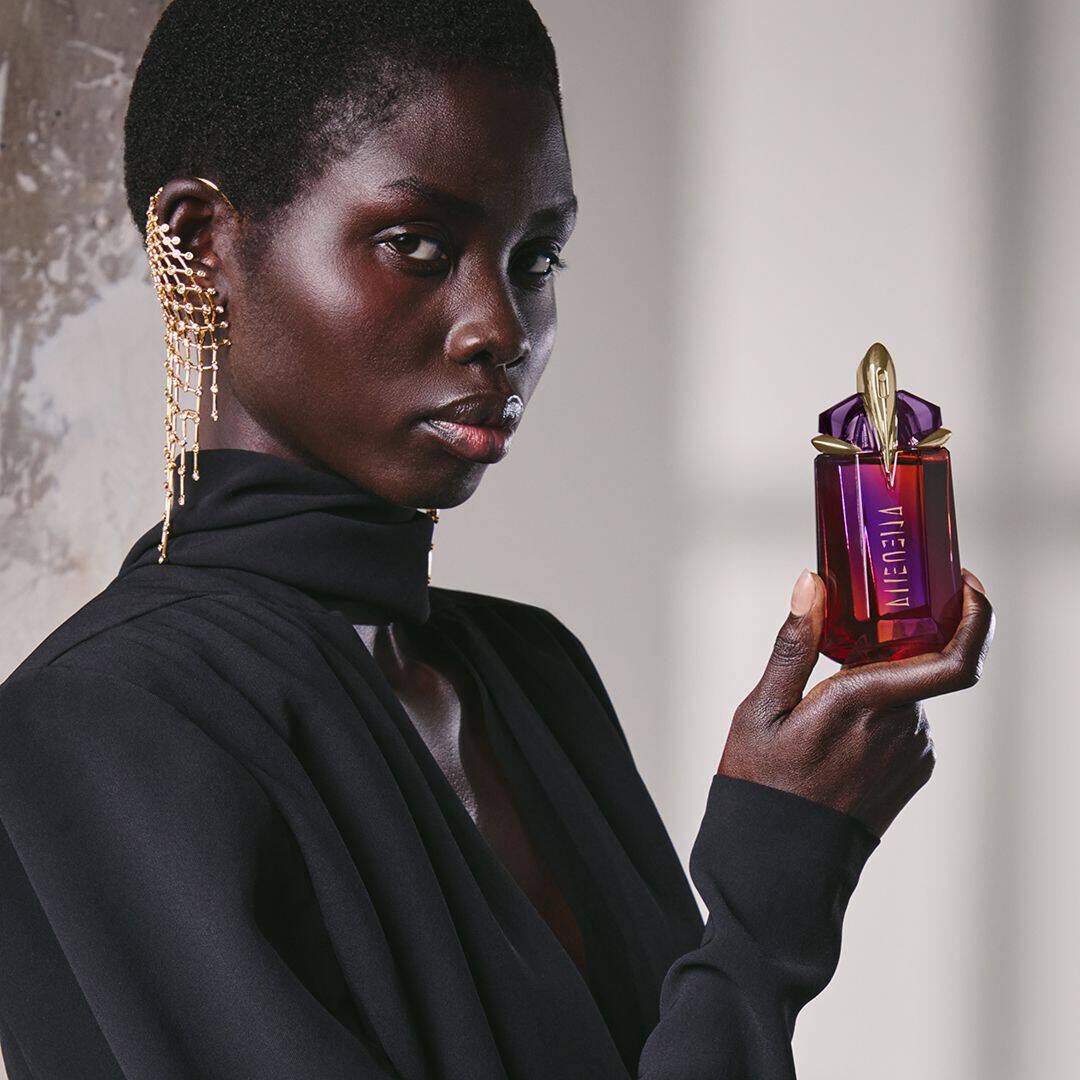 une femme habillee en noir tenant un flacon de parfum rouge et violet dans sa main devant un mur gris
