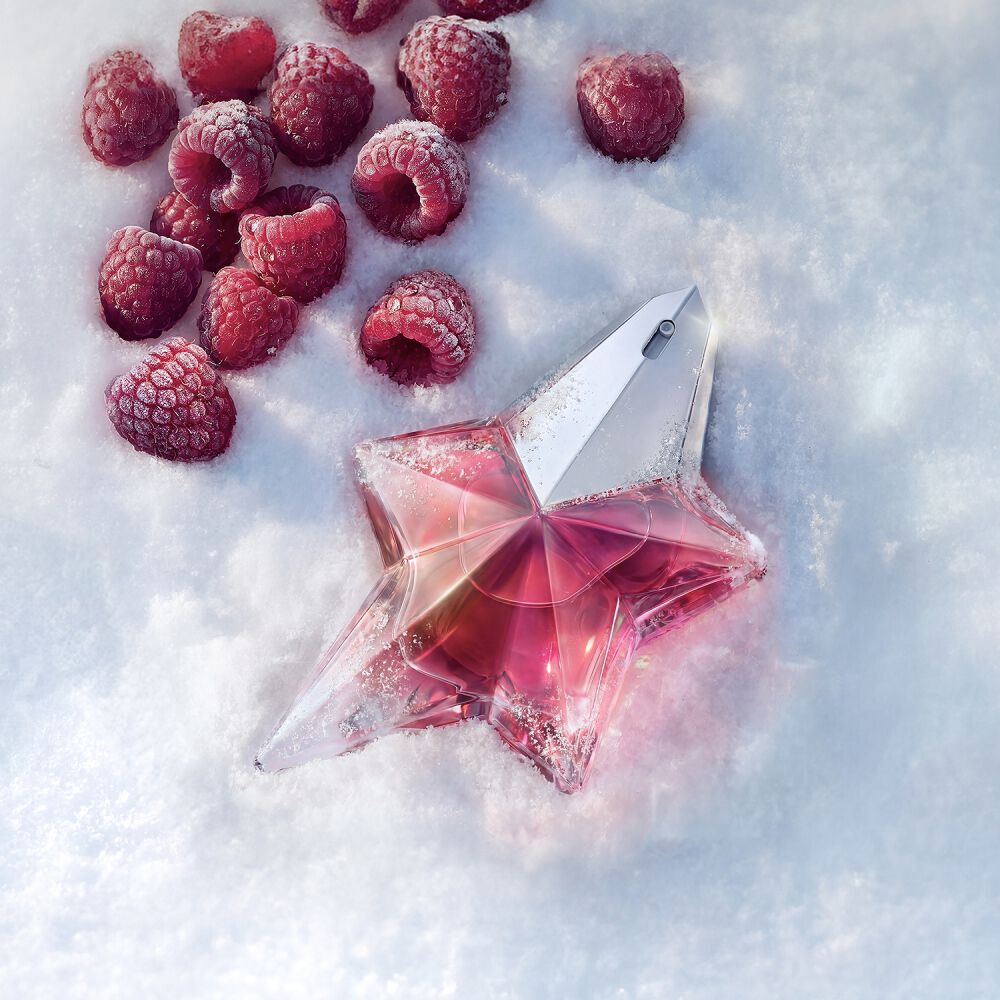 Une bouteille Mugler Angel Nova rose etoile eclatante, posee dans la neige blanche, entouree de framboises rouges fraiches eparpillees qui mettent en valeur ses notes fruitees.