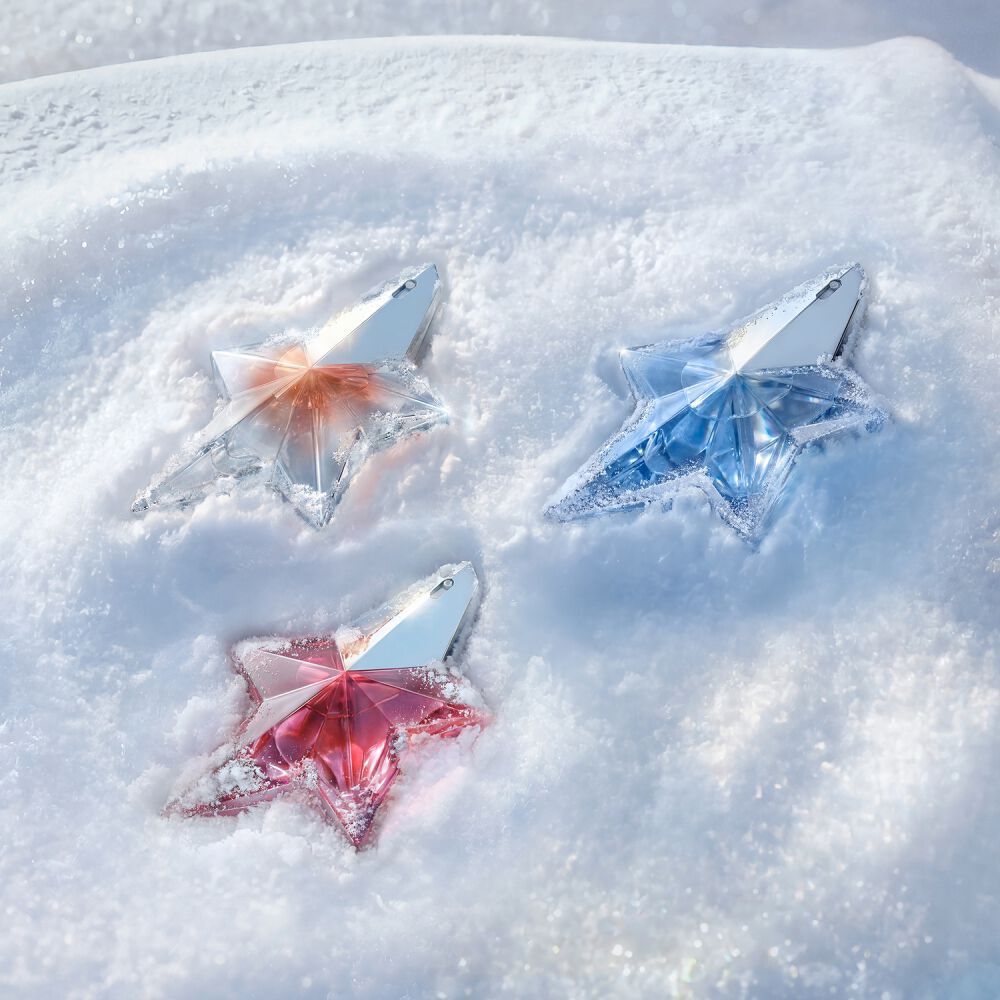 Un trio de flacons de parfums Mugler en forme d’etoile dans des tons d’orange (Angel Blush), bleu clair (Angel eau de parfum) et rose (Angel Nova), disposes ensemble sur une surface neigeuse et etincelante.