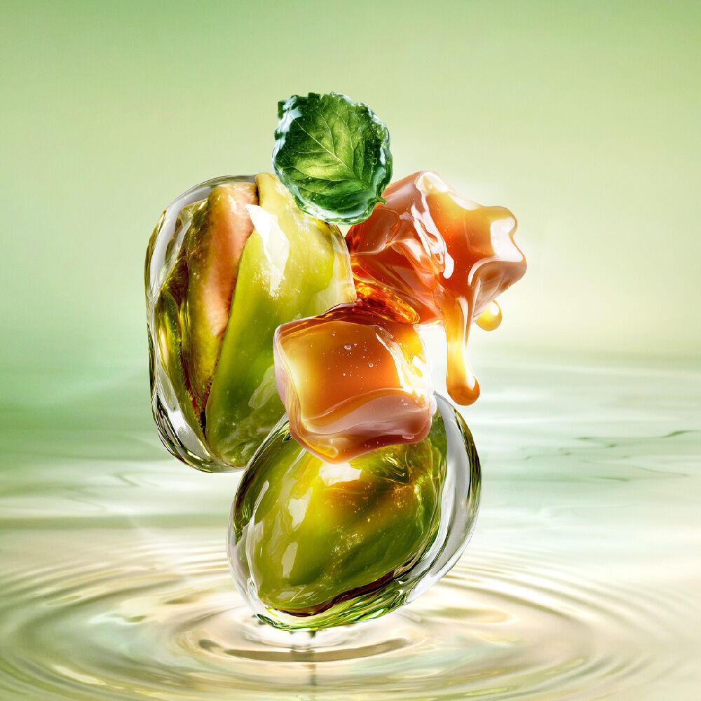 Nouvelle Starlicious Pistachio Praline image de nature morte montrant la fine bouteille entouree de ses ingredients cles : une feuille de patchouli, des pistaches entieres et des cubes de caramel degoulinants, le tout sur un fond d’eau vert lumineux ondulant.