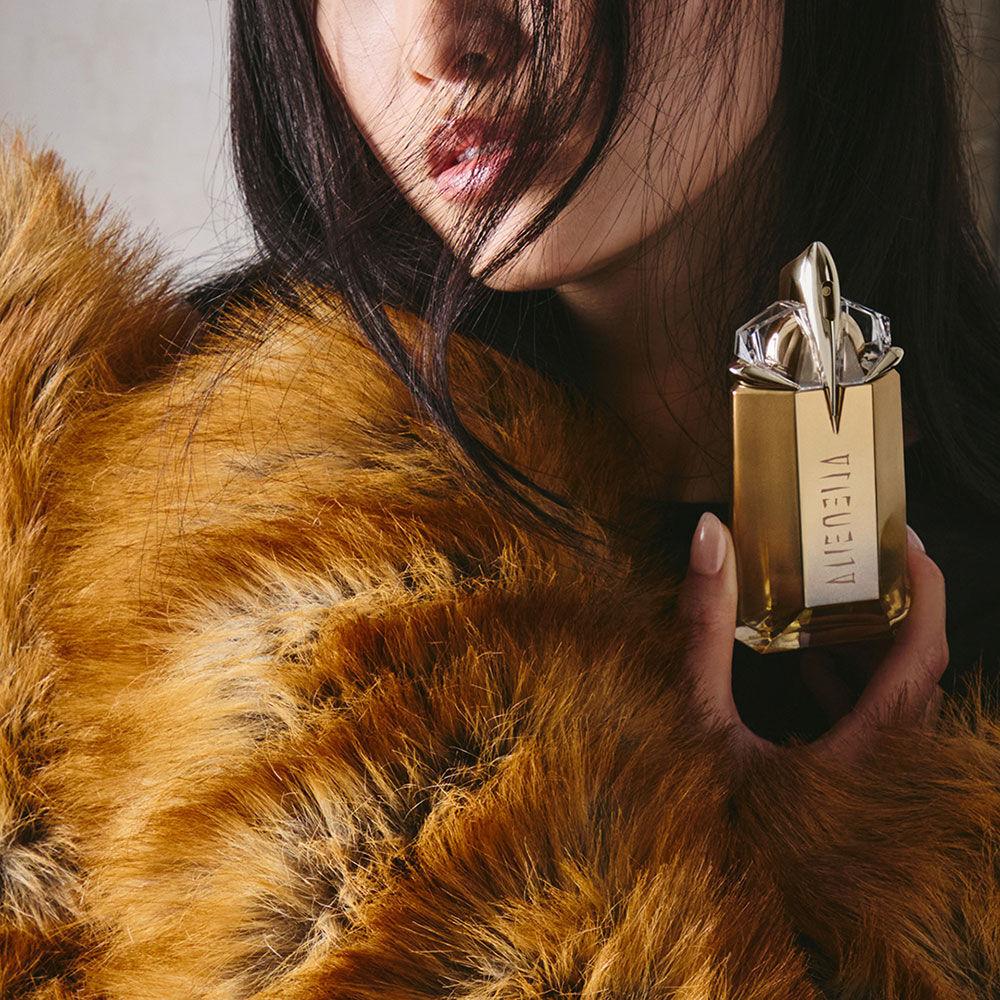 une fois brune portant une fourrure marron tenant dans son main un flacon de parfum dore mugler