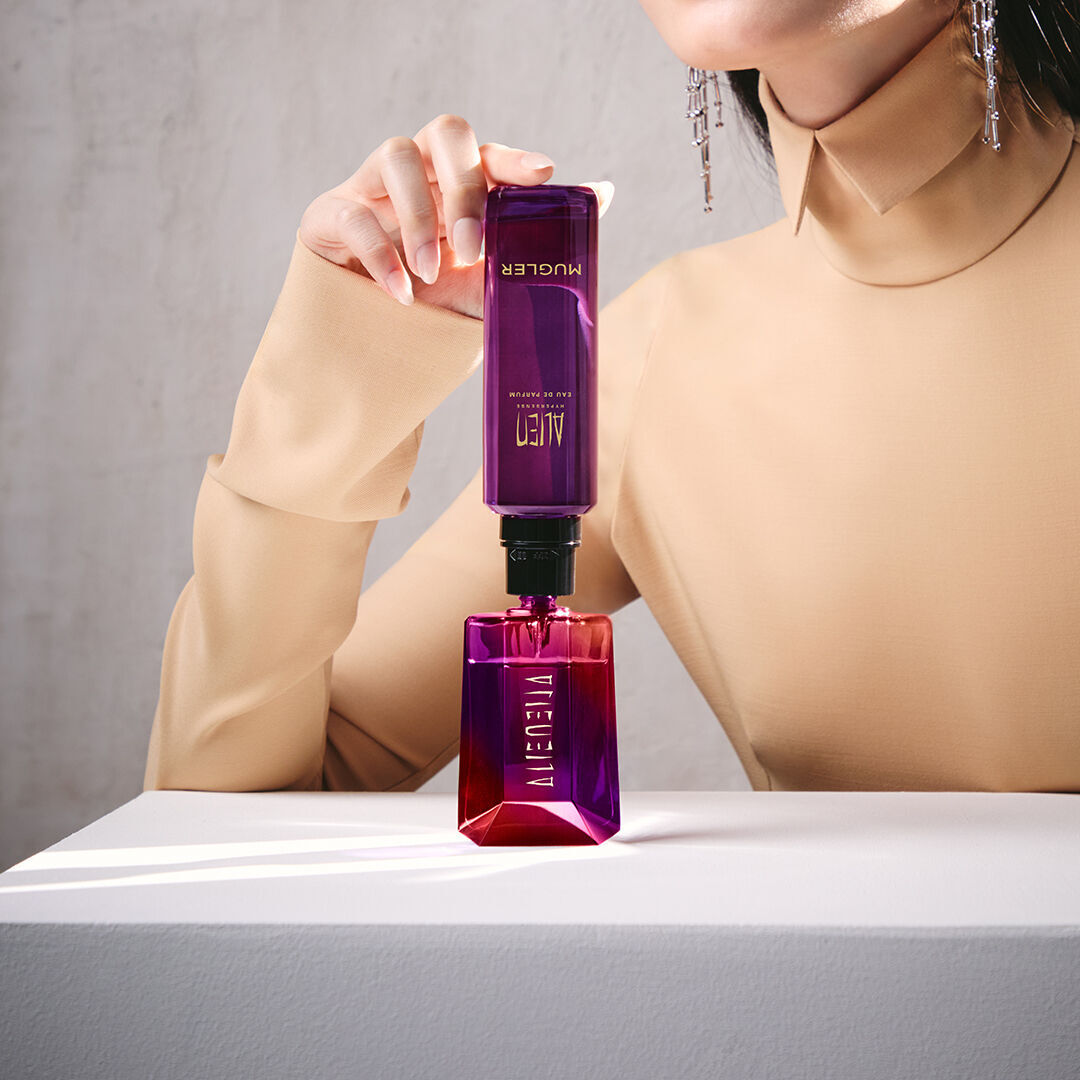 un flacon de parfum rouge et violet remplit par une recharge violette tenu par la main d'une femme habille en beige