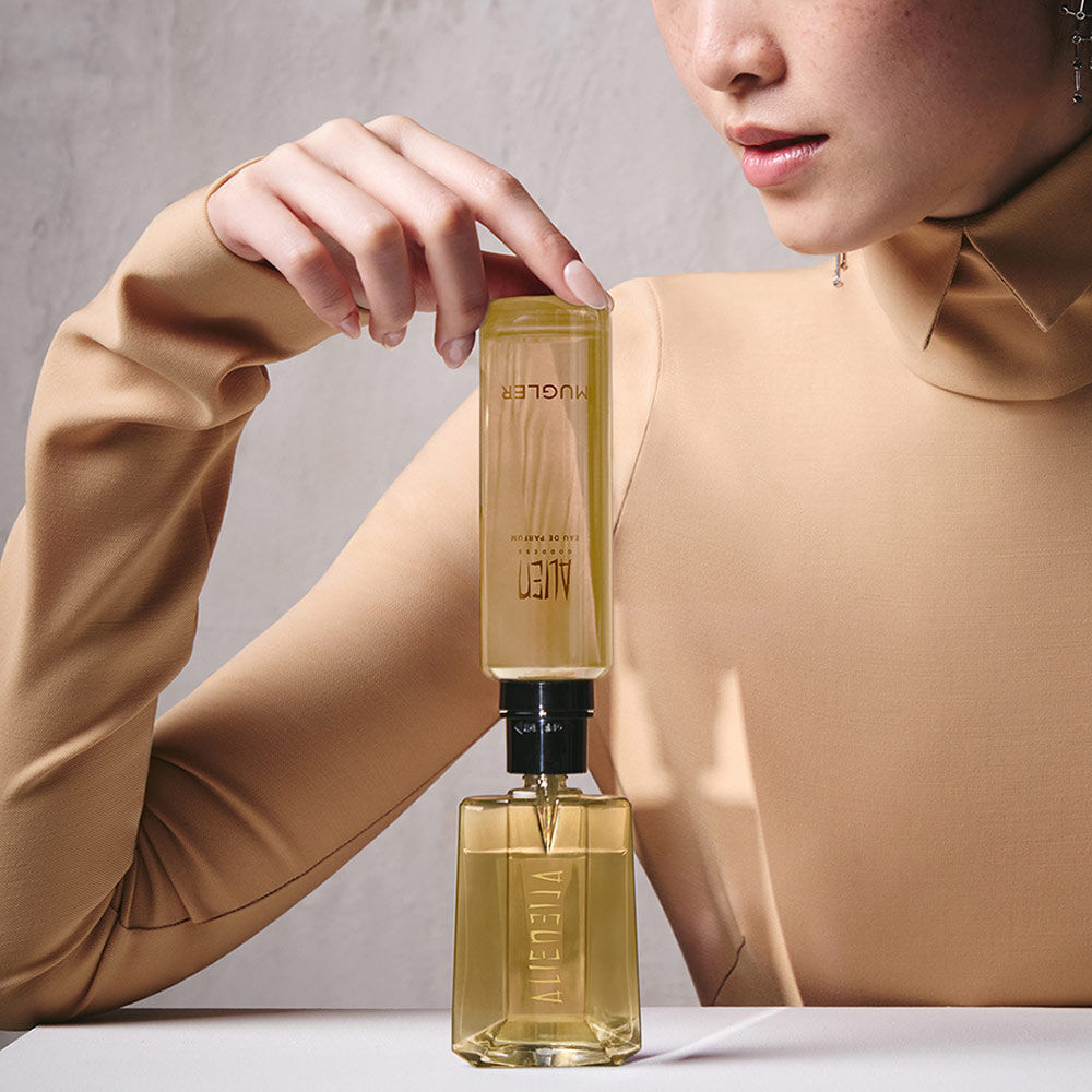 un flacon de parfum dore remplit par une recharge jaune tenu par la main d'une femme habille en beige