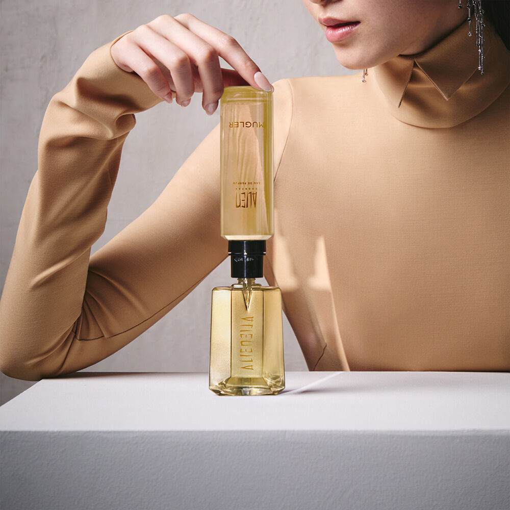 un flacon de parfum dore remplit par une recharge jaune tenu par la main d'une femme habille en beige