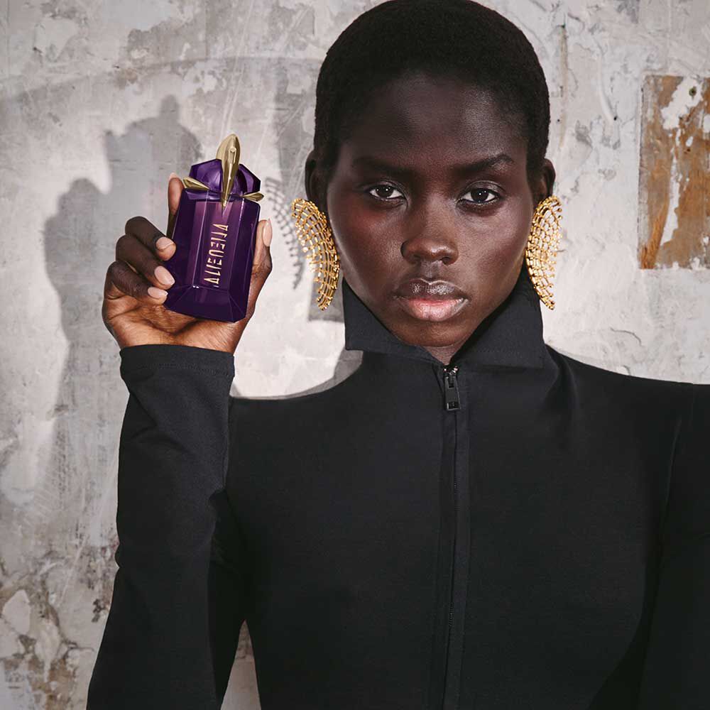 une femme habillee en noir tenant un flacon de parfum violet dans sa main devant un mur gris