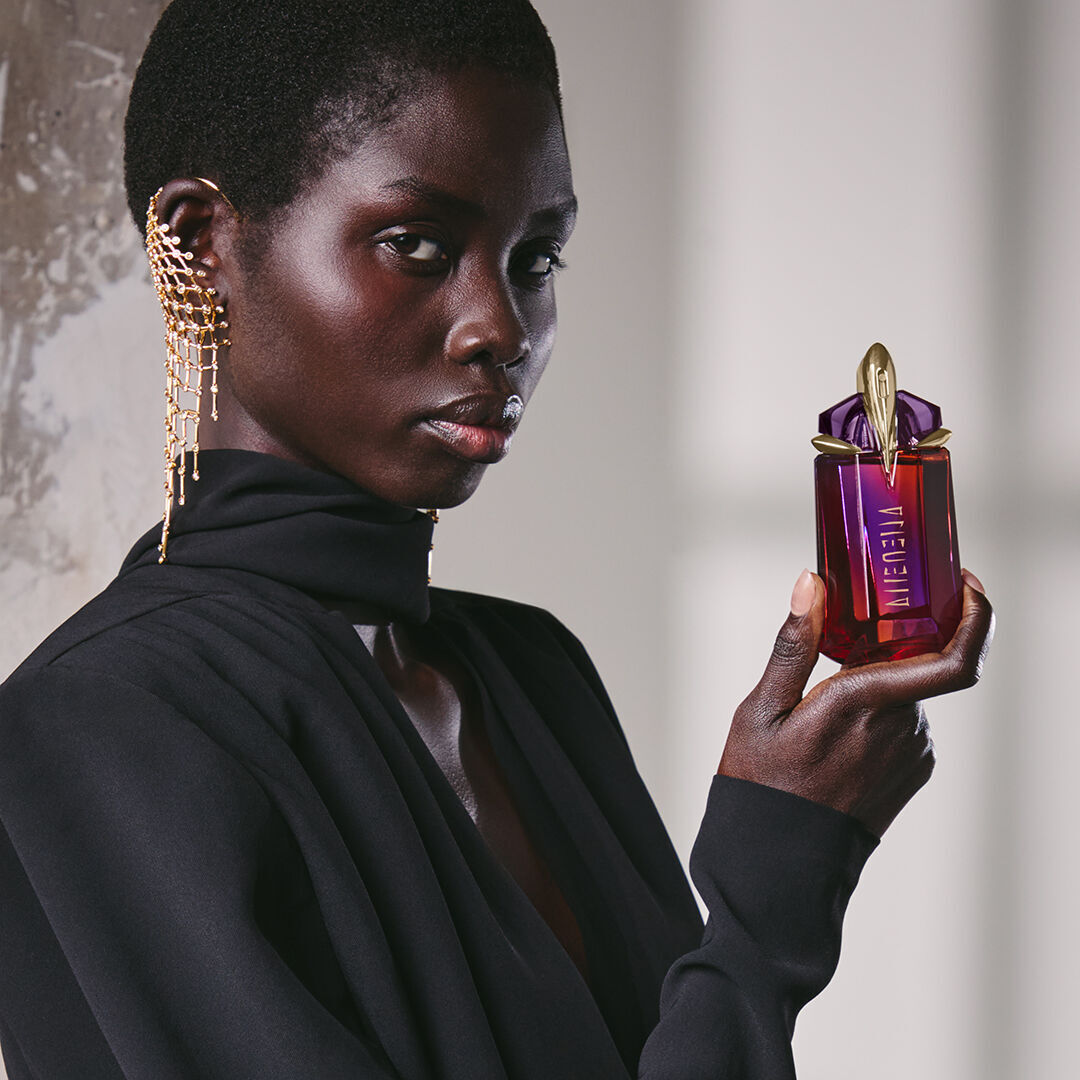 une femme habillee en noir tenant un flacon de parfum rouge et violet dans sa main devant un mur gris