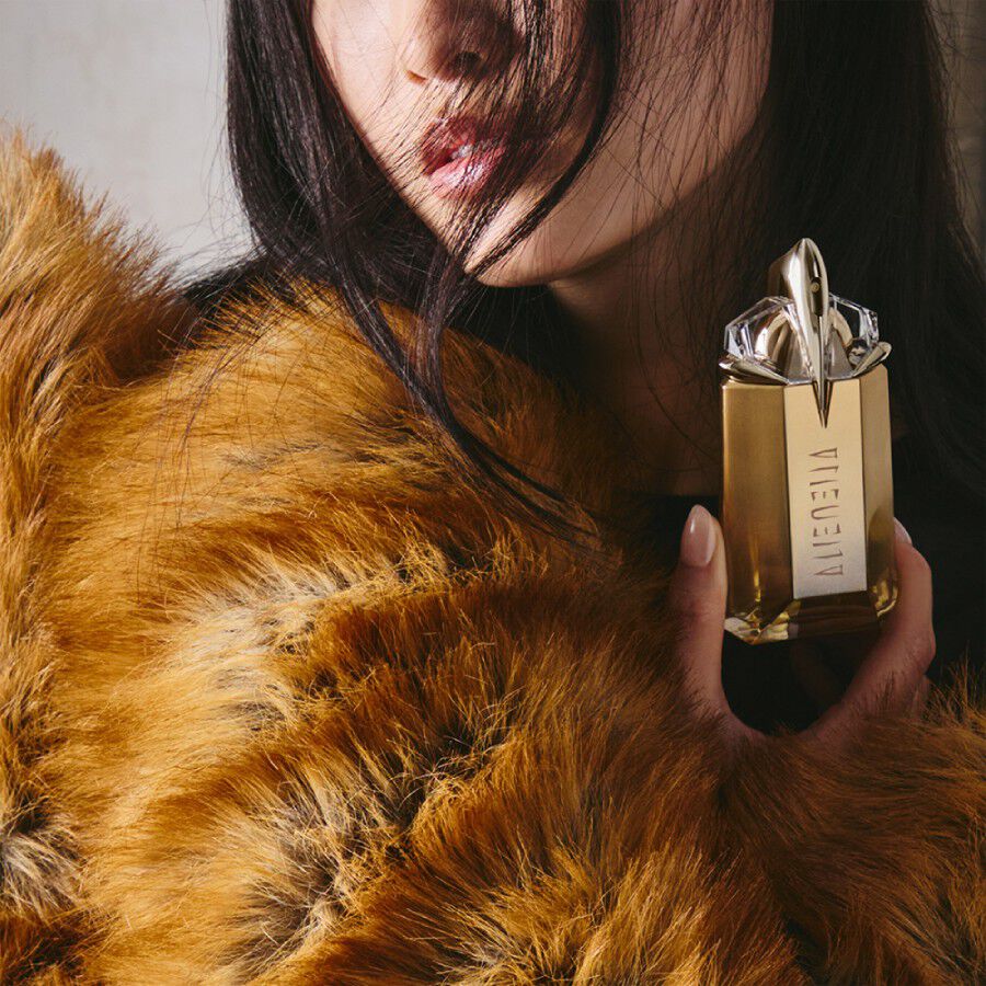 une fois brune portant une fourrure marron tenant dans son main un flacon de parfum dore mugler