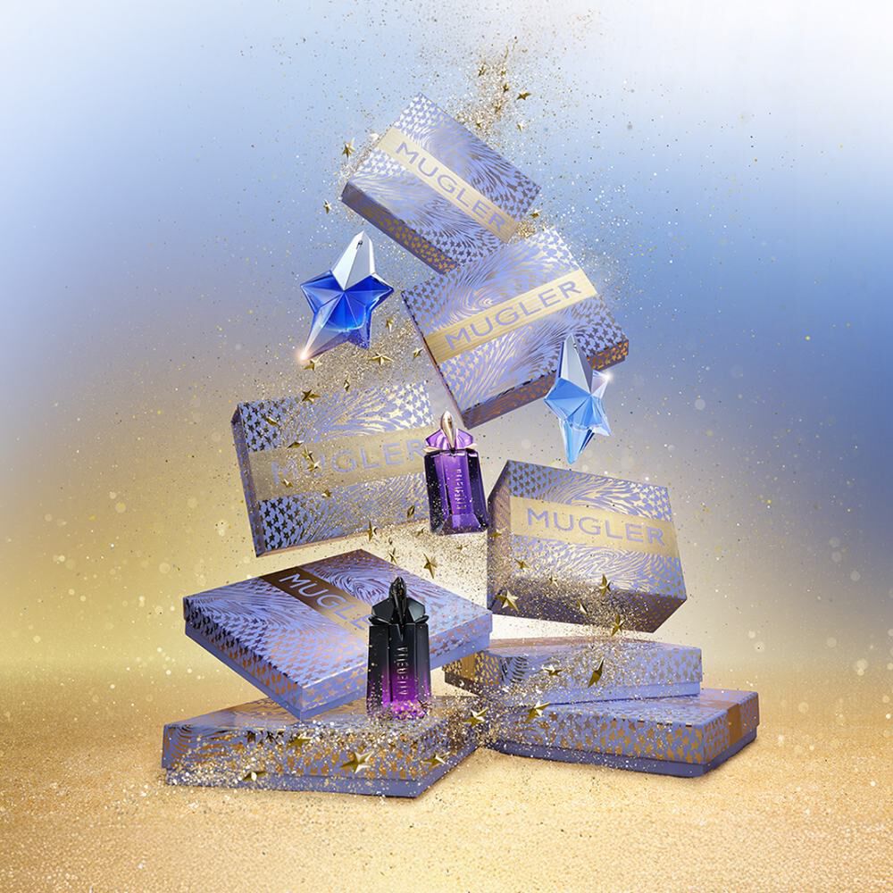 Une composition festive de coffrets cadeaux et de flacons de parfum Mugler de differentes tailles et couleurs (bleu, violet) est disposee. Des etoiles et des paillettes dorees sont dispersees partout, creant une ambiance magique sur un fond degrade bleu et dore.
