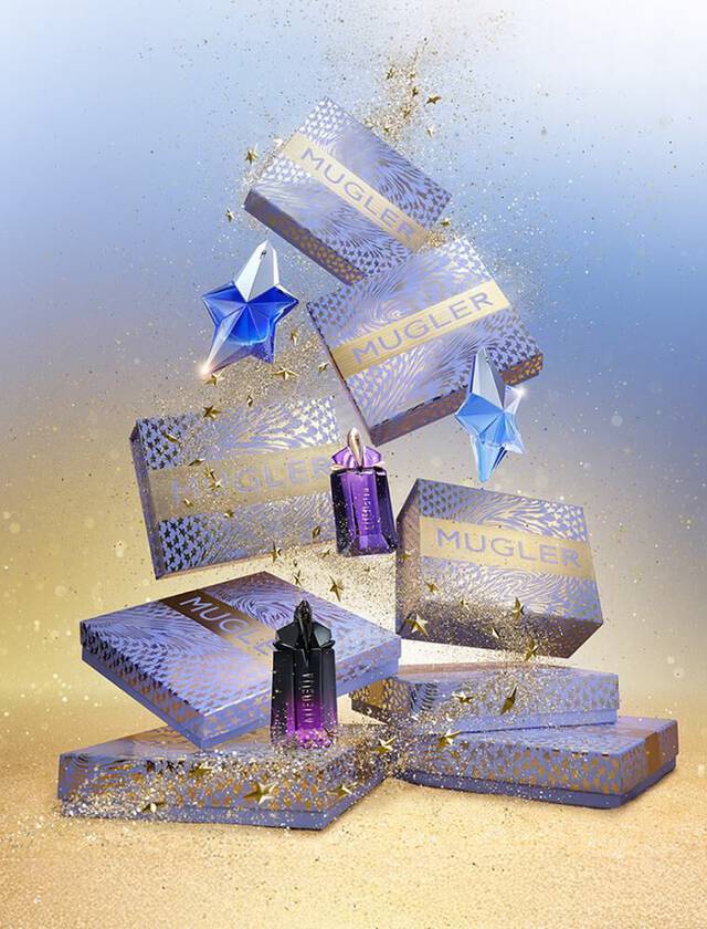 Une composition festive de coffrets cadeaux et de flacons de parfum Mugler de differentes tailles et couleurs (bleu, violet) est disposee. Des etoiles et des paillettes dorees sont dispersees partout, creant une ambiance magique sur un fond degrade bleu et dore.