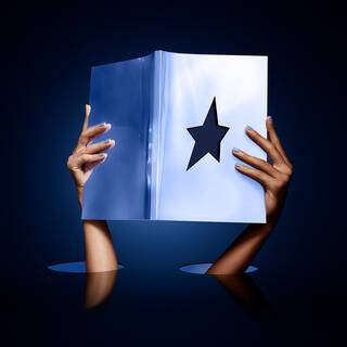 A pair of hands holding a glossy book open to reveal a black star cutout against a dark blue background, with two circular holes beneath the hands.