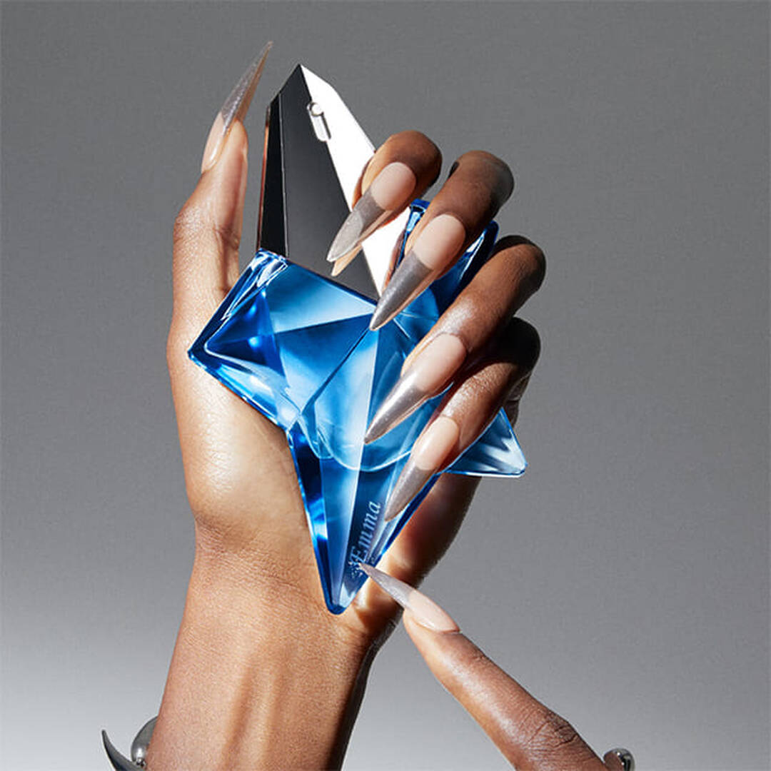 Une main noire avec des ongles longs et pointus tient un flacon de parfum en forme d'etoile, de couleur bleu translucide.