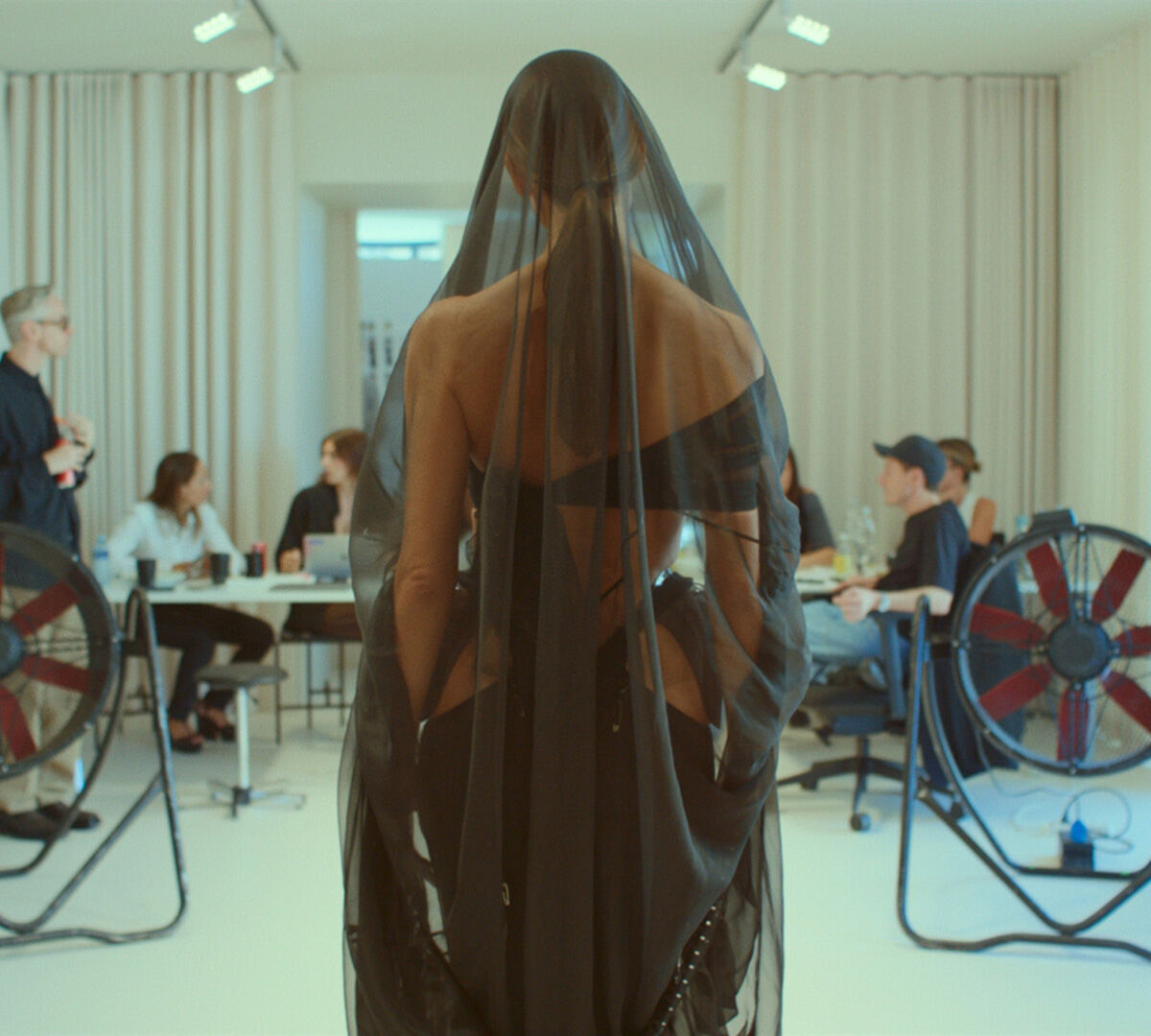 Photographie d'une femme au dos nu, portant un long voile noir transparent, debout dans une piece baignee de lumiere. A l'arriere-plan, plusieurs personnes sont assises autour d'une table, et deux grands ventilateurs industriels sont visibles.