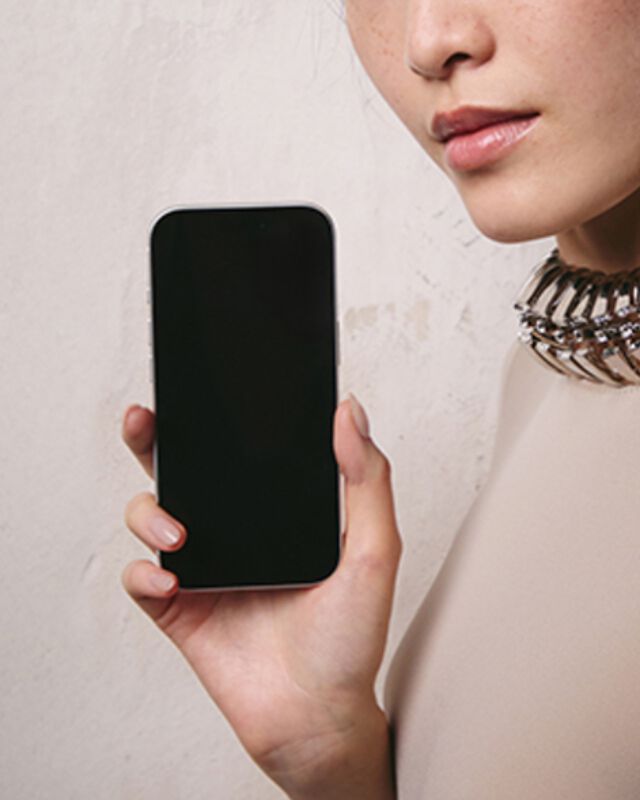Une femme aux cheveux fonces portant un haut de couleur claire et un collier a col metallique, tenant un smartphone avec un ecran vide, devant un mur gris clair texture.