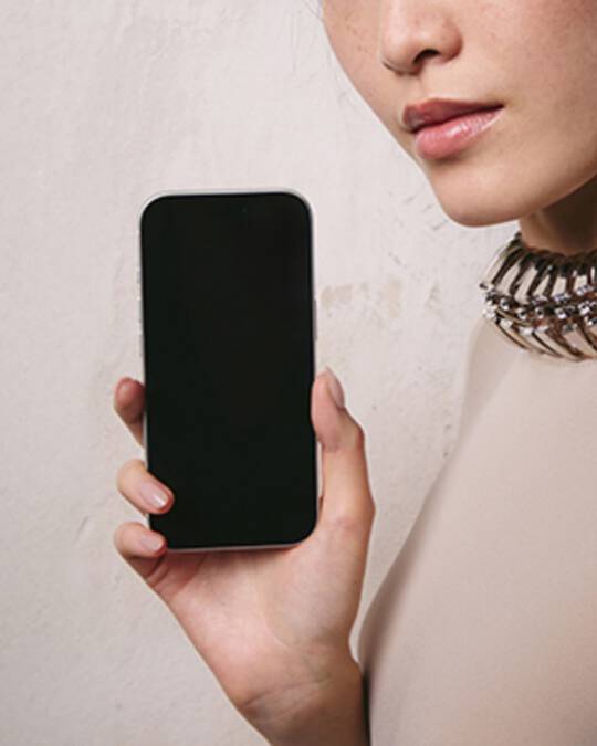 Une femme aux cheveux fonces portant un haut de couleur claire et un collier a col metallique, tenant un smartphone avec un ecran vide, devant un mur gris clair texture.
