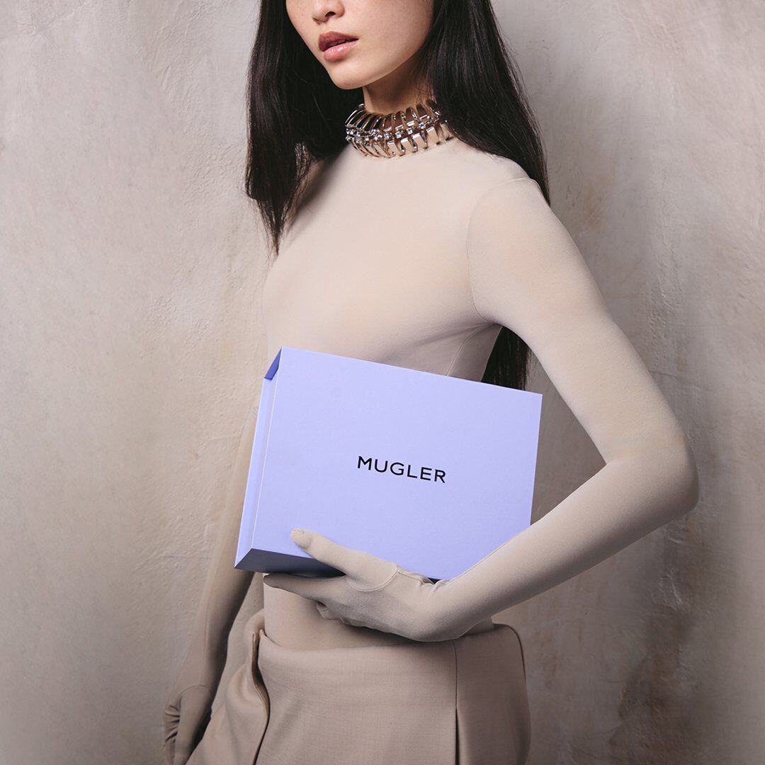 Mannequin vetue d'un col roule beige et d'un collier ras-du-cou argente, tenant un coffret cadeau Mugler couleur lavande devant un mur gris