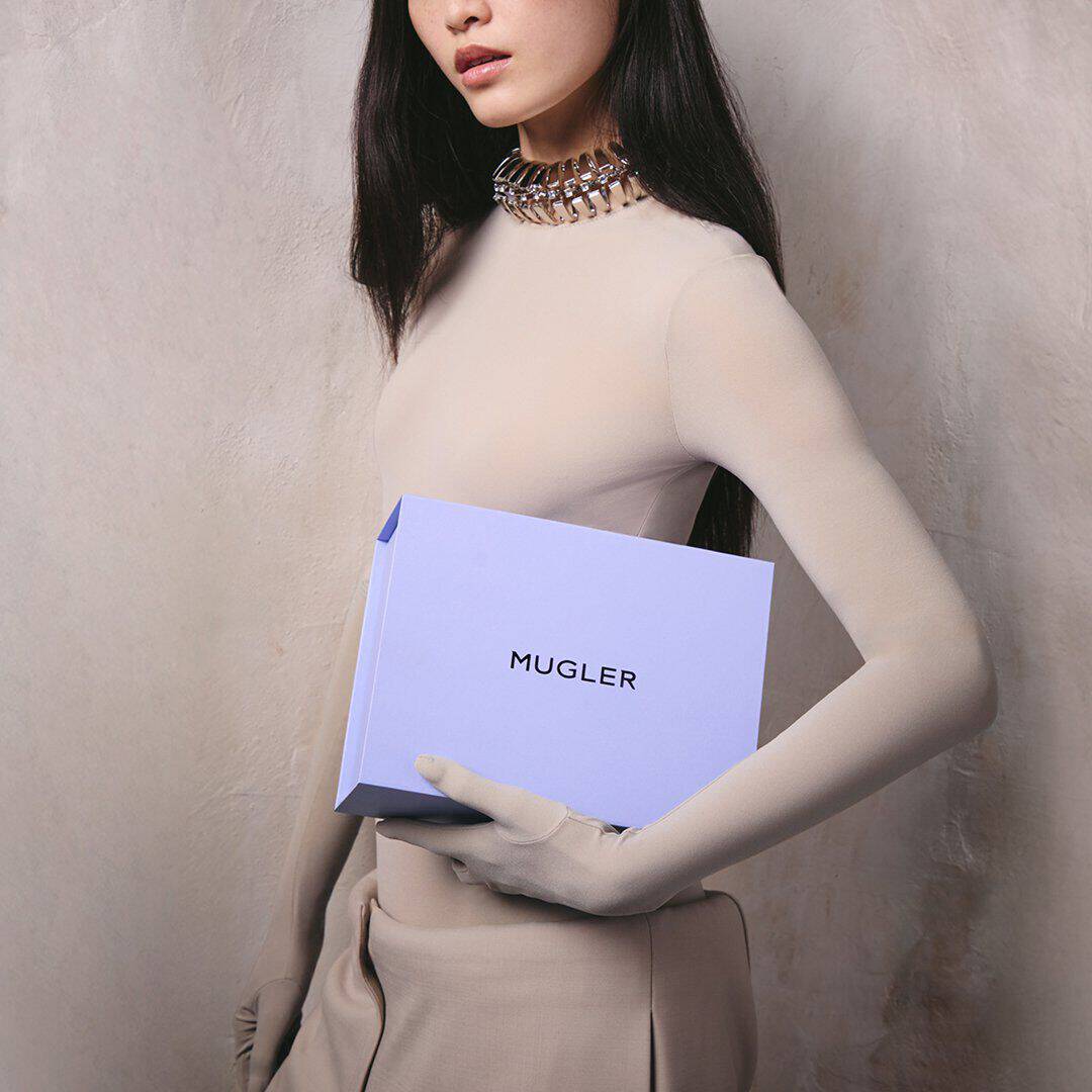 Mannequin vetue d'un col roule beige et d'un collier ras-du-cou argente, tenant un coffret cadeau Mugler couleur lavande devant un mur gris