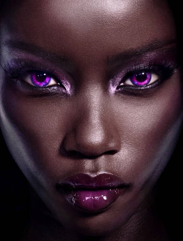 Portrait d'une personne avec un teint sombre, mettant en valeur des yeux violets brillants et un maquillage saisissant, sur un fond noir.