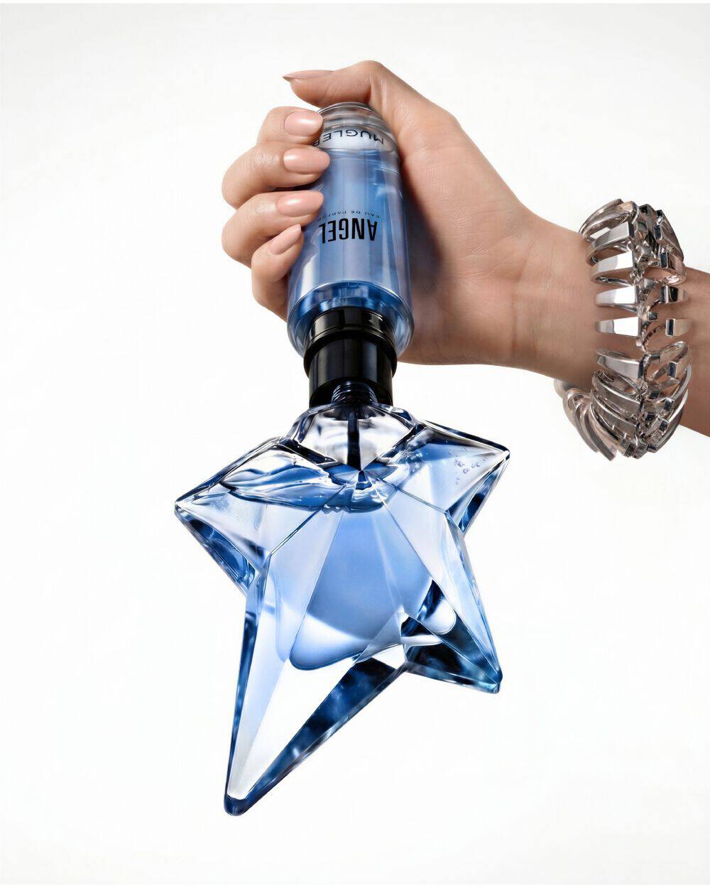 Gros plan sur une main en train de remplir le flacon emblematique bleu en forme d'etoile de l'eau de parfum « Angel » a l'aide d'un flacon de recharge Mugler bleu, sur un fond beige clair laissant entrevoir une partie du visage d'un mannequin.