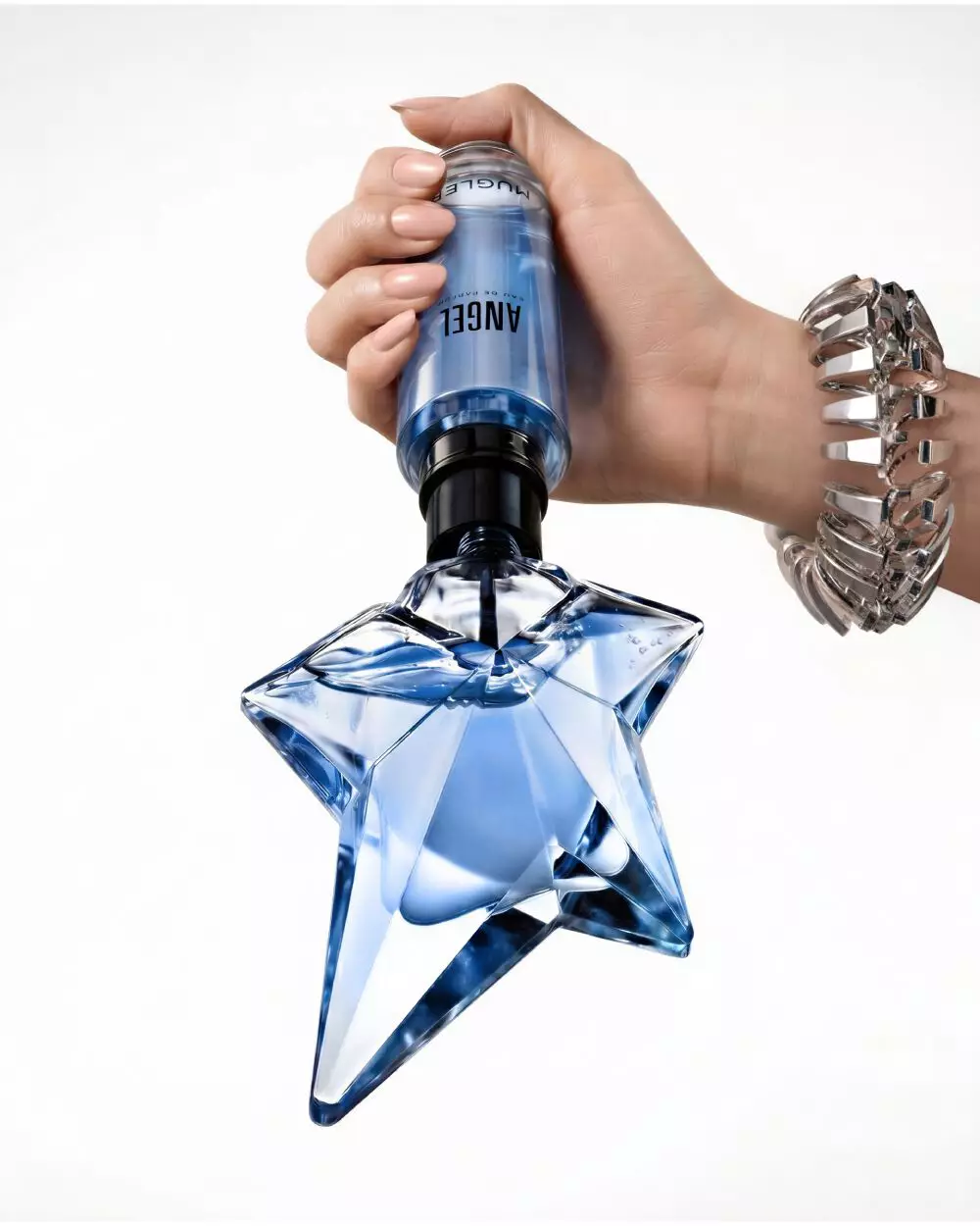 Gros plan sur une main en train de remplir le flacon emblematique bleu en forme d'etoile de l'eau de parfum « Angel » a l'aide d'un flacon de recharge Mugler bleu, sur un fond beige clair laissant entrevoir une partie du visage d'un mannequin.