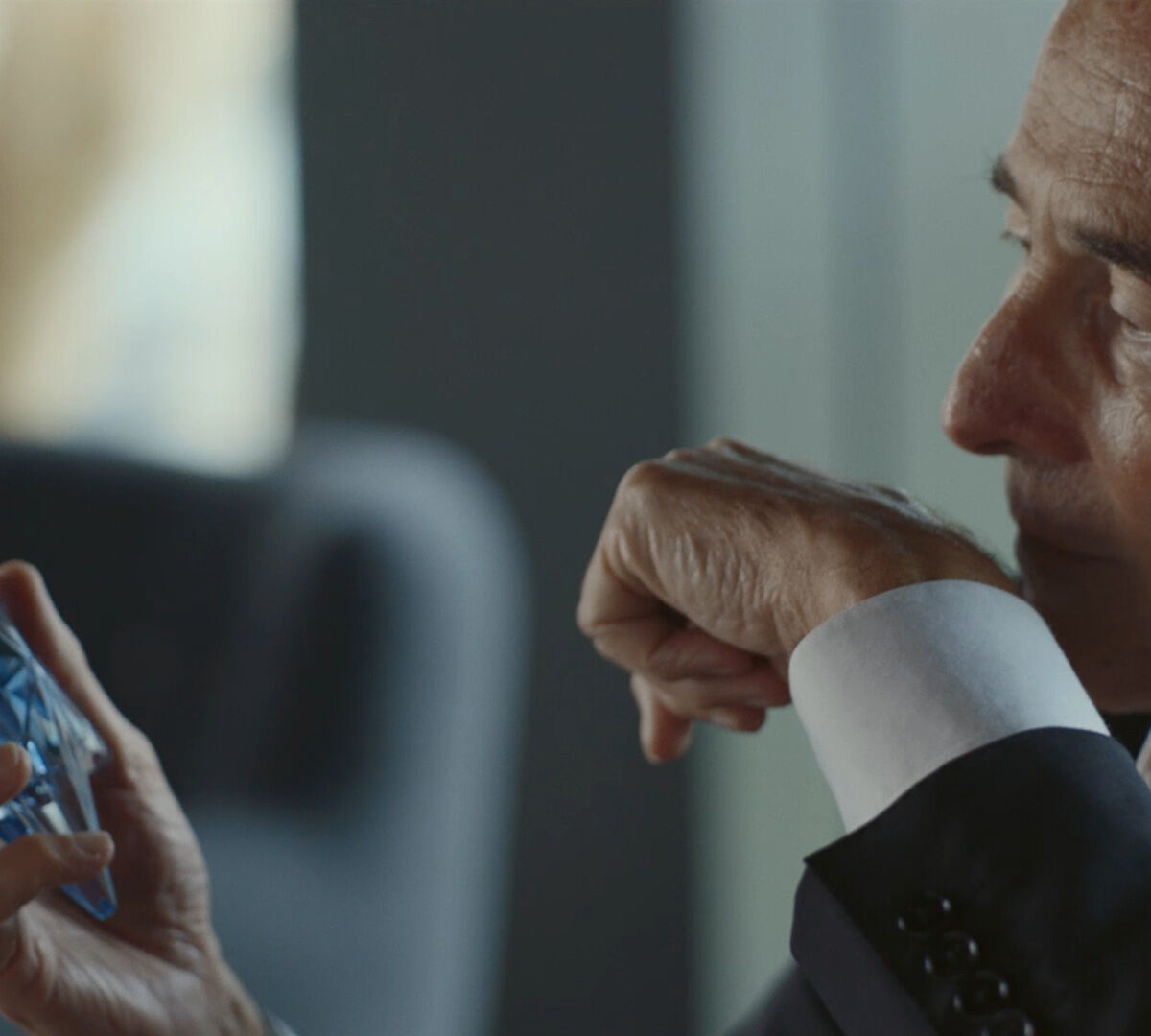 Gros plan sur un homme age en costume, tenant un flacon de parfum bleu dans sa main gauche, tandis que sa main droite est levee pres de son visage