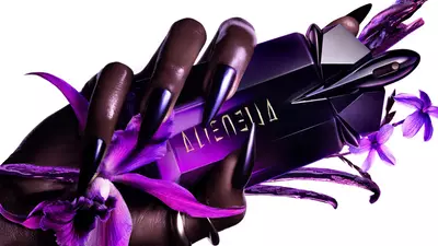Un flacon de parfum aux teintes violettes, tenu par une main avec de longs ongles noirs, entoure de fleurs violettes.