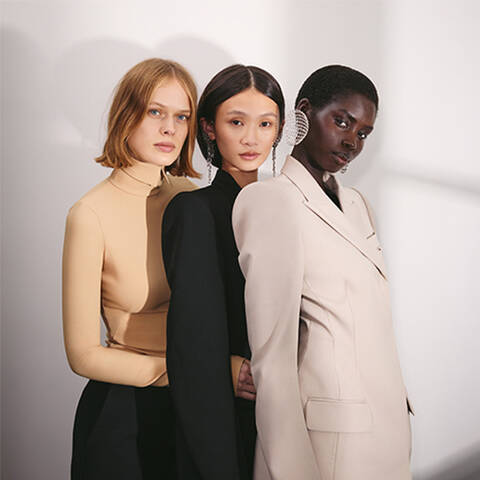 Three women standing together, facing forward, one in a tan turtleneck, one in a black blazer with long earrings, and one in a light beige blazer with large, intricate hoop earrings