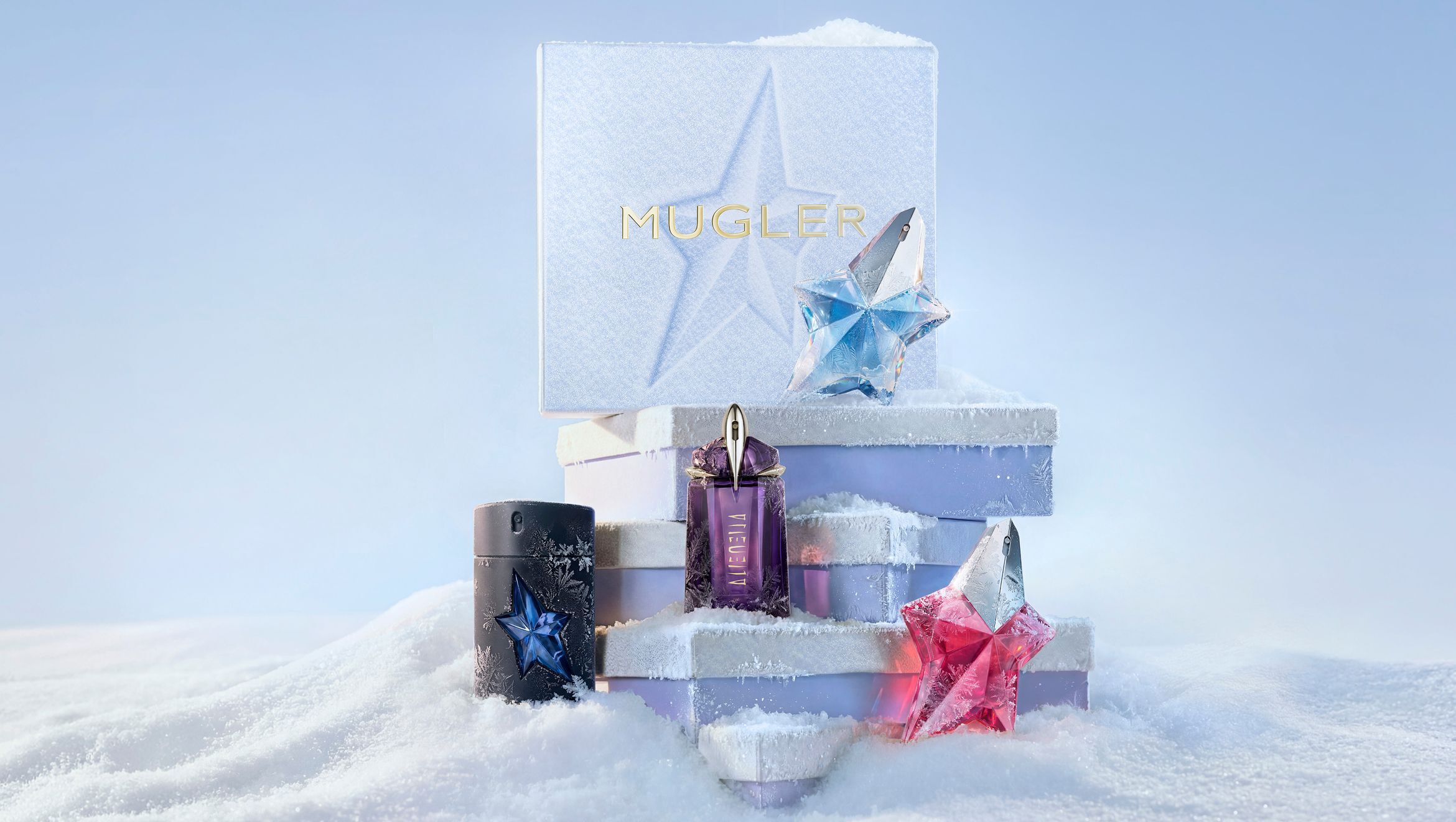 A stack of three light blue Mugler gift sets piled above the snow surrounded by a black A*Men eau de toilette bottle, a purple Alien eau de parfum bottle, a blue Angel eau de parfum bottle, and a pink Angel eau de parfum bottle in a snowy landscape.