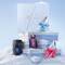 A stack of three light blue Mugler gift sets piled above the snow surrounded by a black A*Men eau de toilette bottle, a purple Alien eau de parfum bottle, a blue Angel eau de parfum bottle, and a pink Angel eau de parfum bottle in a snowy landscape.