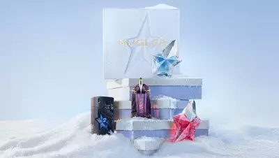A stack of three light blue Mugler gift sets piled above the snow surrounded by a black A*Men eau de toilette bottle, a purple Alien eau de parfum bottle, a blue Angel eau de parfum bottle, and a pink Angel eau de parfum bottle in a snowy landscape.