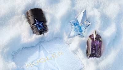 A black A*Men eau de toilette bottle, a blue star-shaped Angel eau de parfum bottle, and a purple Alien eau de parfum bottle lie together in the snow next to a glistening holiday gift set.