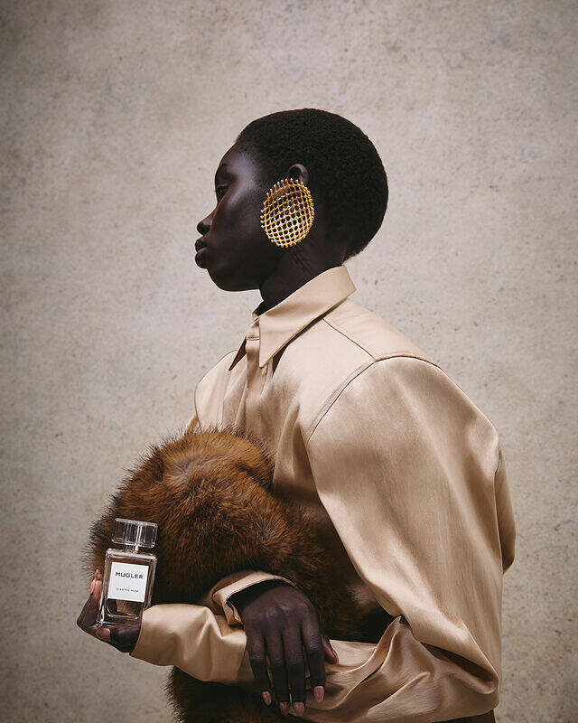 Gros plan sur une main tenant le parfum « Mugler Over The Musk » devant un fond de fourrure luxueux.