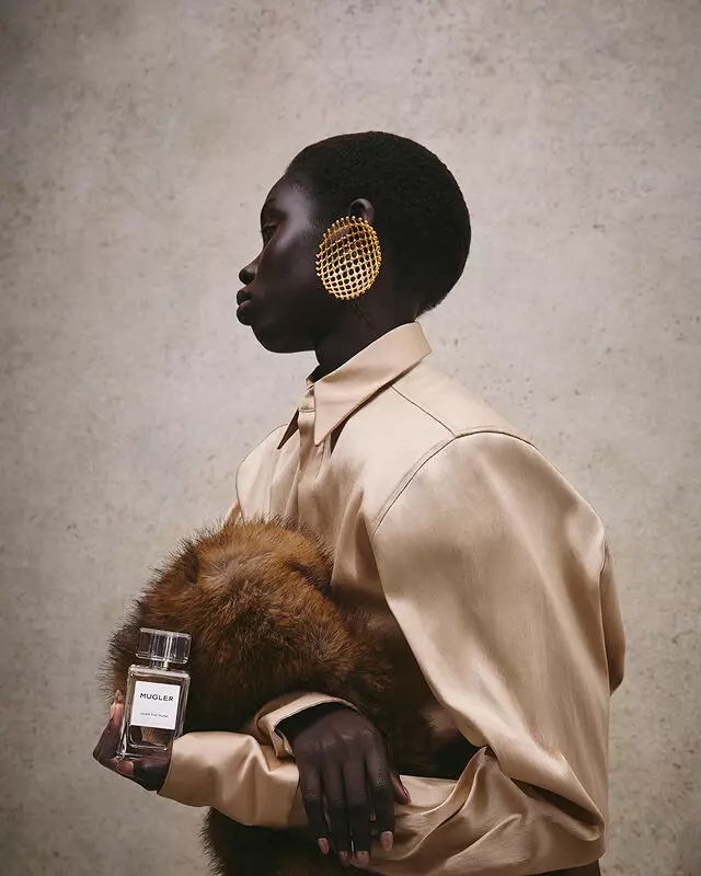 Gros plan sur une main tenant le parfum « Mugler Over The Musk » devant un fond de fourrure luxueux.