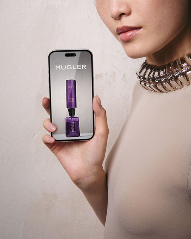 Une femme aux cheveux fonces portant un haut de couleur claire et un collier a col metallique, tenant un smartphone avec un ecran vide, devant un mur gris clair texture.