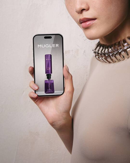 Une femme aux cheveux fonces portant un haut de couleur claire et un collier a col metallique, tenant un smartphone avec un ecran vide, devant un mur gris clair texture.