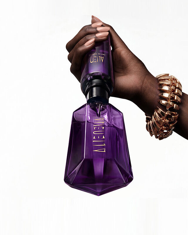 Une main portant un bracelet en maillons d'or en train de recharger chez elle un flacon violet d'Eau de Parfum Alien a l'aide d'un flacon de recharge violet officiel, sur lequel figure la mention « rechargez chez vous ».