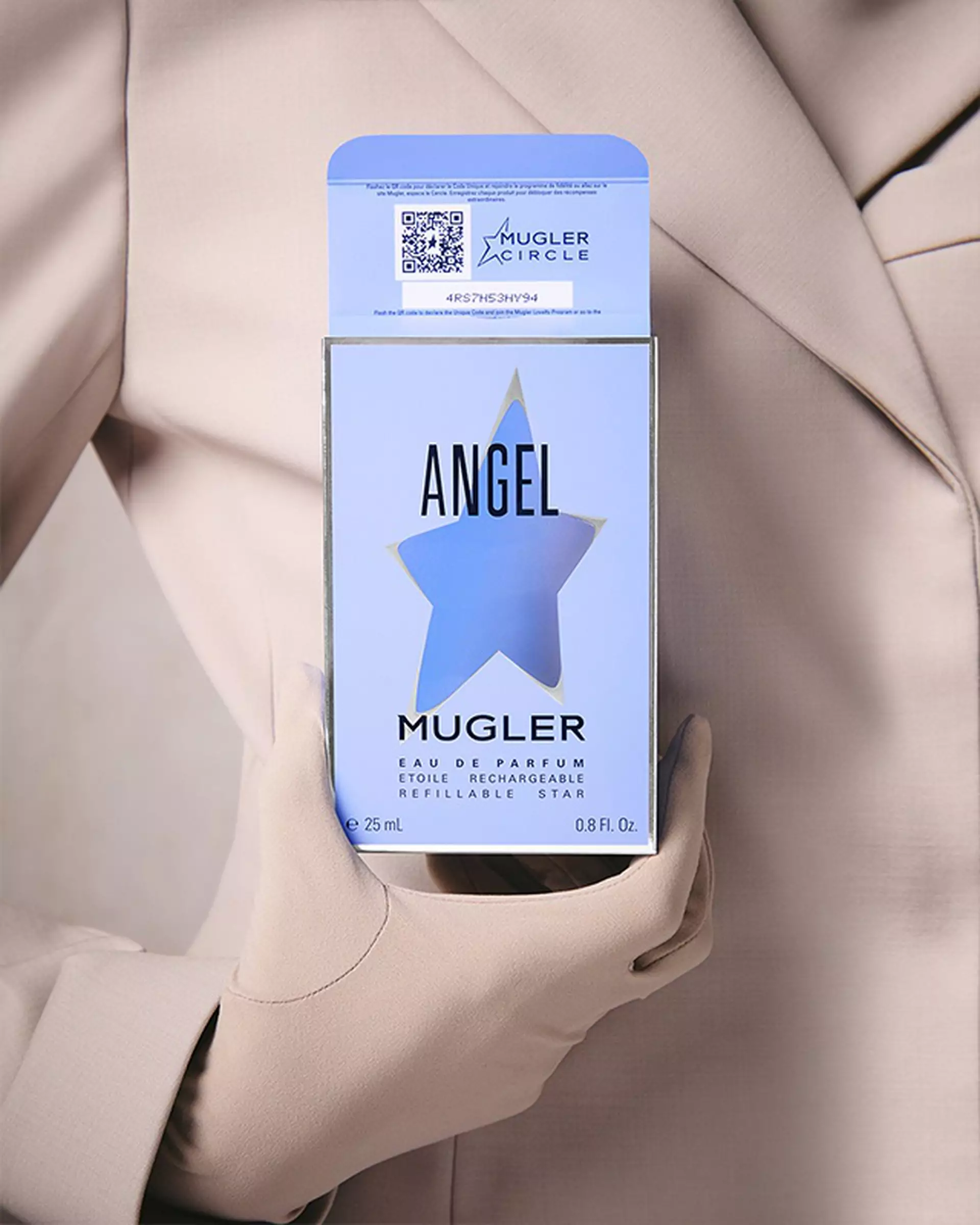 Une personne vetue d'un blazer et de gants beige clair, tenant une boite de parfum bleu clair avec une decoupe en forme d'etoile et du texte noir.