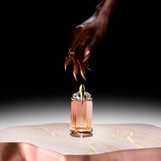 Main elegante avec des ongles longs tenant un flacon de parfum sur une surface marbree. Le fond est sombre, mettant en valeur le flacon.
