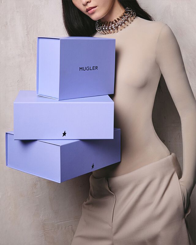 Une personne vetue d'un haut et d'un pantalon beiges, tenant trois boites lavande empilees ornees d'etoiles noires, un collier metallique sur la poitrine nue, sur un fond neutre texture.