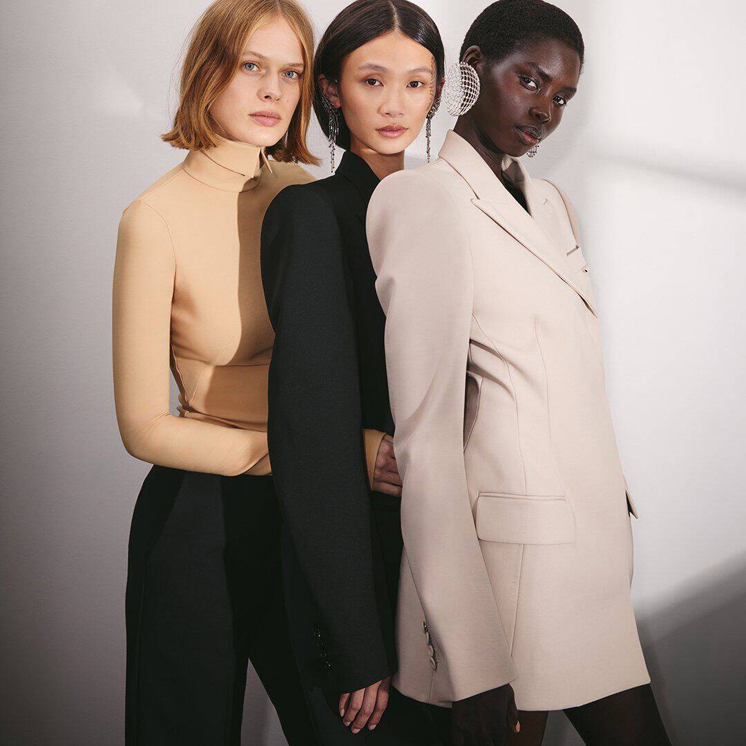 Trois mannequins vetues de tenues beiges et noires, arborant des boucles d'oreilles imposantes, posent ensemble devant un fond neutre