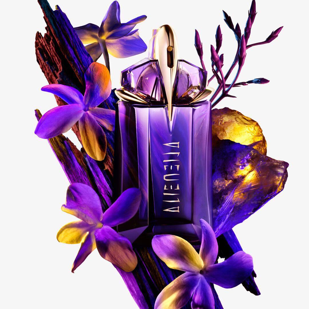 Une bouteille de parfum violette entouree de fleurs et de cristaux, sur un fond clair.