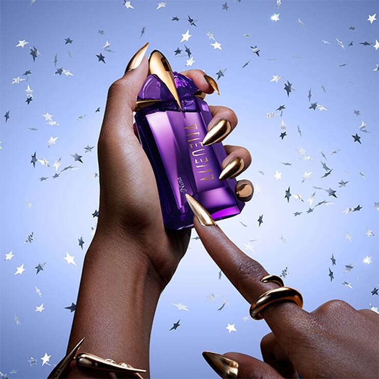 Une main avec des ongles dores tient un flacon de parfum violet, tandis qu'un doigt touche le flacon, sur un fond bleu parseme d'etoiles argentees.