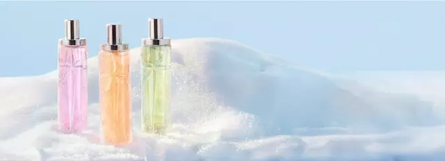 Trois grands flacons de parfum Mugler de forme cylindrique, aux teintes translucides de rose, d'orange et de vert clair, alignes debout dans la neige. La nouvelle collection Starlicious.