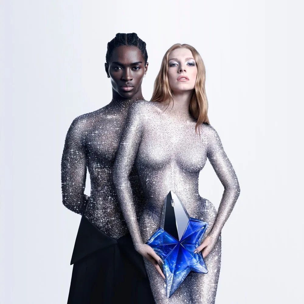 Un homme et une femme, vetus de tenues chatoyantes, regardent la camera. Elle tient une etoile dans ses mains, evoquant l'un des parfums de Mugler.
