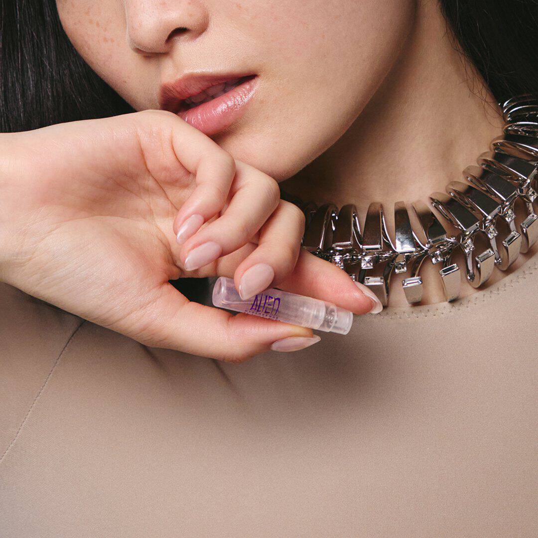 Gros plan sur un mannequin portant un collier a chaine argentee et tenant pres de son visage un flacon d'echantillon du parfum « Alien » de Mugler