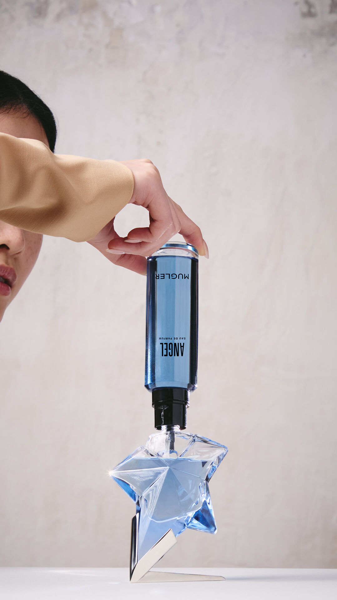 Une main en train de remplir le flacon bleu en forme d'etoile de l'Eau de Parfum « Angel » a l'aide d'un flacon de recharge bleu place a l'envers.