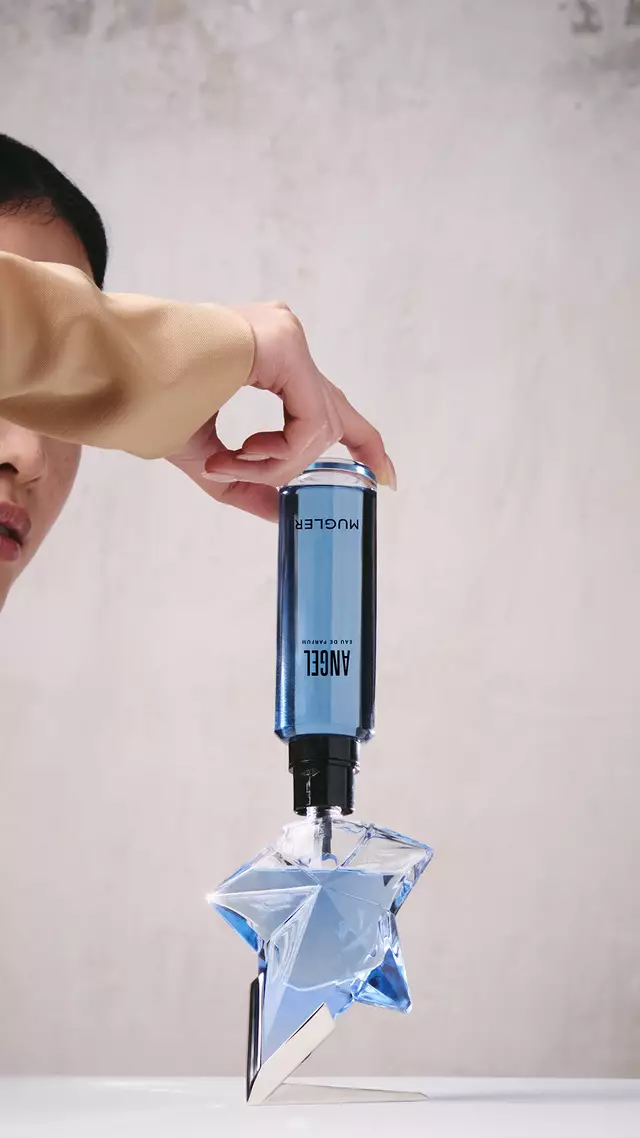 Une main en train de remplir le flacon bleu en forme d'etoile de l'Eau de Parfum « Angel » a l'aide d'un flacon de recharge bleu place a l'envers.