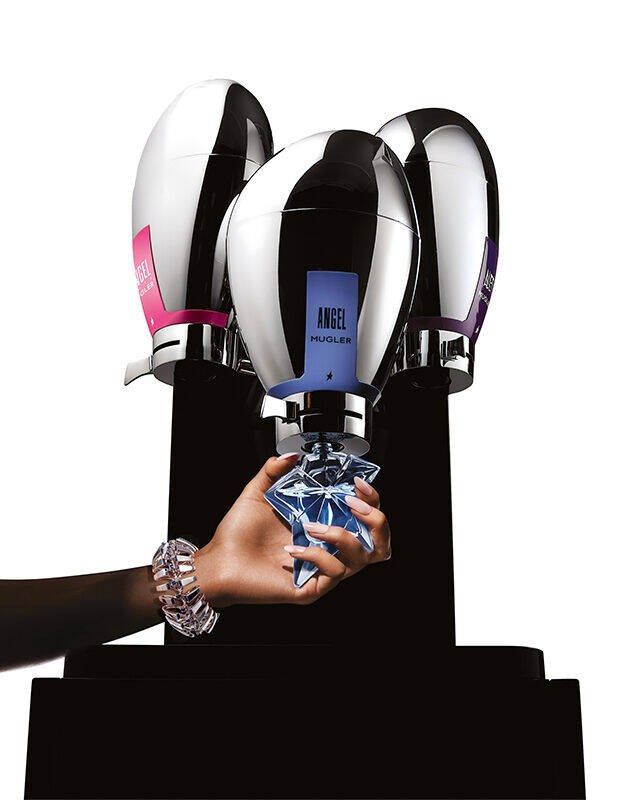 Une fontaine a parfum Mugler au design epure et futuriste, de couleur metallique, dotee de trois buses, avec une main en train de remplir une bouteille en verre placee en dessous.