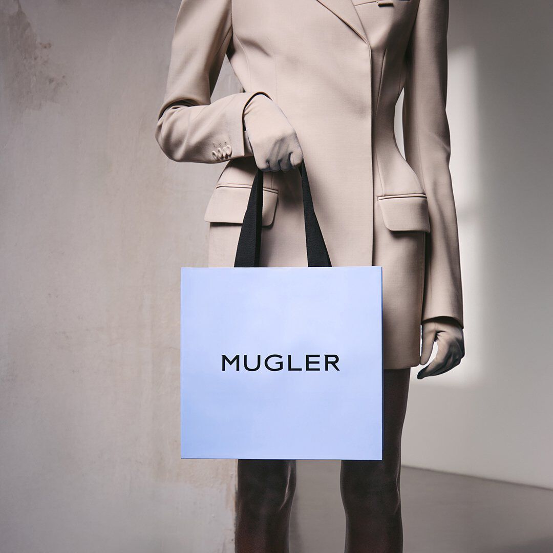 Une personne vetue d'un blazer beige, tenant un sac de courses Mugler bleu clair a anses noires, devant un mur de couleur neutre
