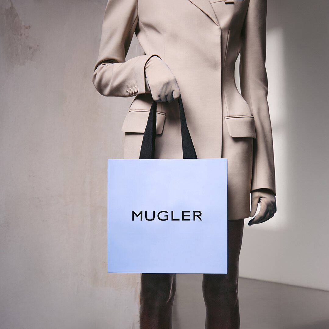 Une personne vetue d'un blazer beige, tenant un sac de courses Mugler bleu clair a anses noires, devant un mur de couleur neutre