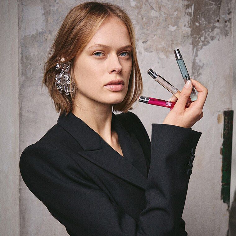 Gros plan sur un mannequin portant une boucle d'oreille argentee a laquelle sont accroches trois flacons de voyage des parfums Mugler, dont Angel et Alien, devant un mur industriel texture.